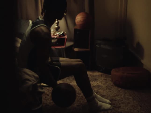 A young athlete sitting in a dimly lit room with a basketball, deep in thought and preparation, reflects on his journey and dreams of future success in sports.