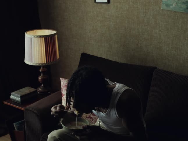 A young black man in a white tank top sits on a couch in a dimly lit room, eating from a bowl