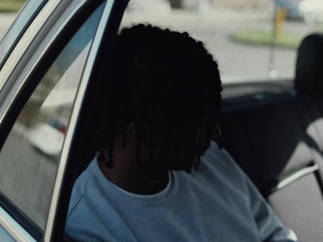 A black teenager with dreadlocks is sitting in the backseat of a car riding through an urban area