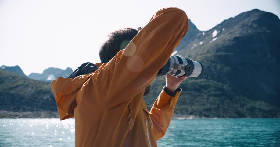 Bearded photographer standing on bobbing surface across from mountainous coast uses telephoto lens to take picture