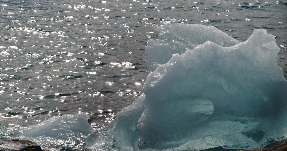 An Iceberg on a rocky shoreline melting into the water with waves reflecting the sun