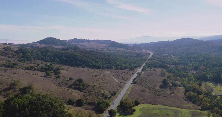 Aerial over a raod through a rural green hillside during the day with mountains in the background