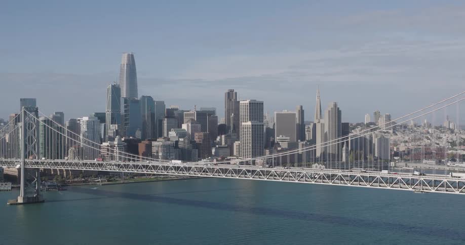 Aerial of suspension bridge in front of a matching silver and gray toned city skyline with blue water and blue sky during the day