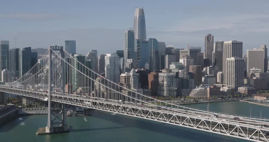 A large bridge with vehicles that goes over the water and goes to a metropolitan city with tall buildings