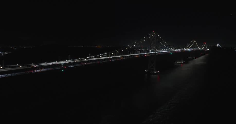 Low aerial over the bay with lights from a long cable suspension bridge reflecting off the black water at night