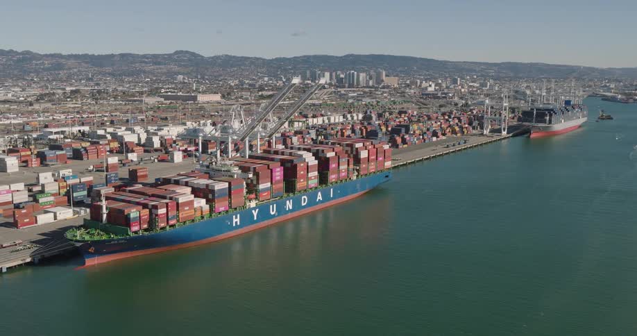Drone aerial of a cargo container stacked ship docked beside a shipping yard under a crane on a bright day