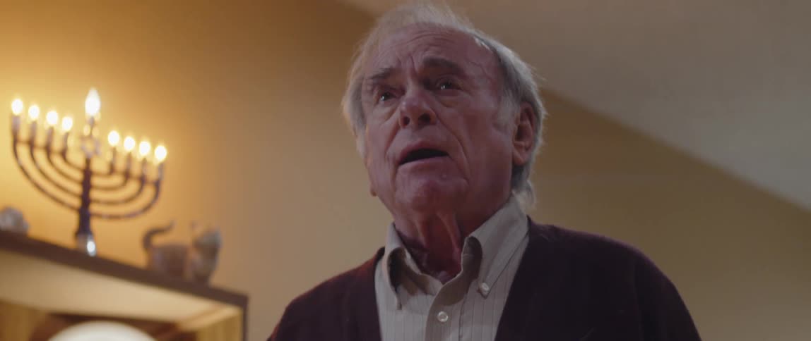An elderly man has a book in his hand and is talking while there is a candlestick lit in front of him and on a shelf behind him