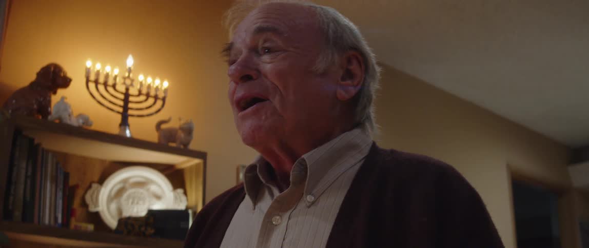 An old man is in a room and is talking and behind him there is a candlestick with lit candles