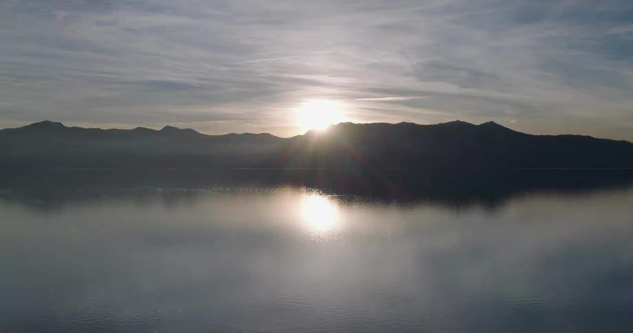 Sun crests silhouetted mountains and shines onto reflecting water partially obscured by fog