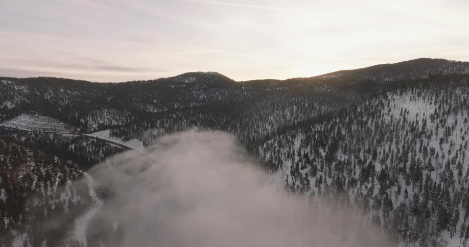 Aerial over a fog covered valley between snow and pine tree covered hills silhouetted by the sunlight rising up from behind mountain peaks on the horizon