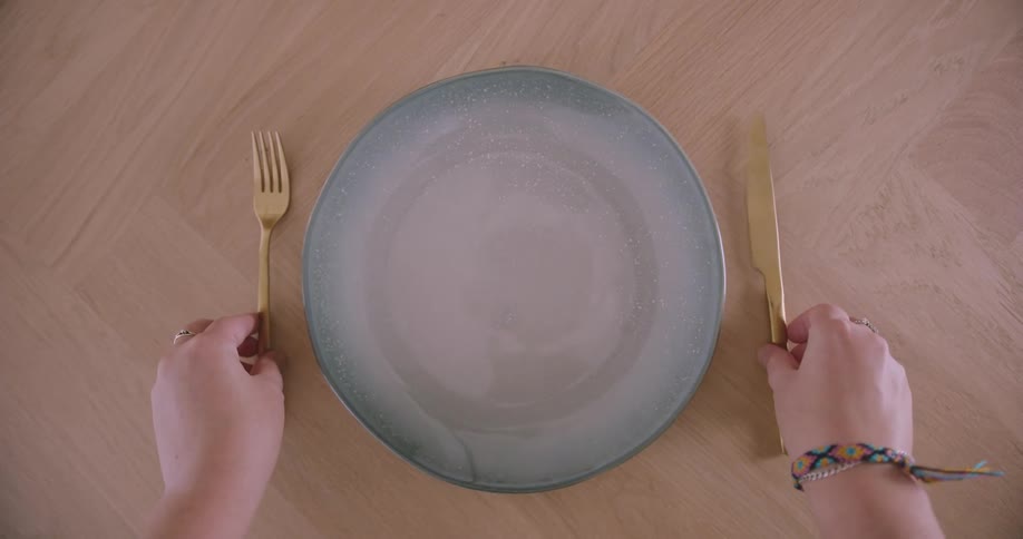 An individual arranges a dining table placing a plate fork knife and cup in preparation for a meal