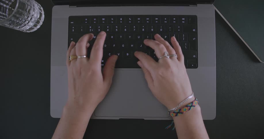 A laptop computer sits on a table as a persons fingers are seen moving on the touch pad and typing on the keyboard
