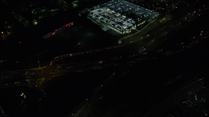 Aerial view headlights moving along two crisscrossed highways beside a white led lit up parking garage at night