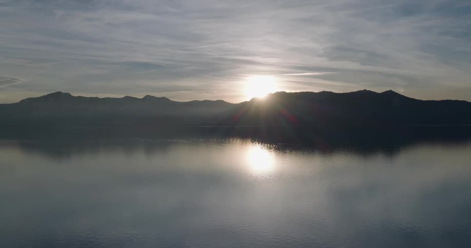 The sun shines through clouds over mountains reflecting on the water along the coast