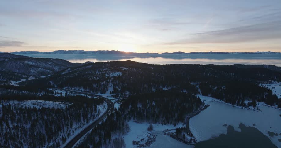 Aerial of a snow covered forest by a single road surrounded by a lake and mountains at sunset