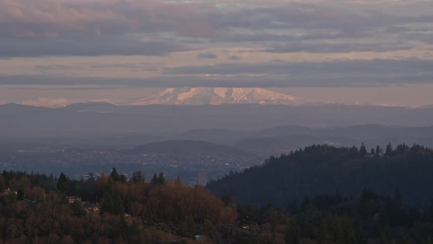 Aerial of a tree covered hillside and city in the valley below with a large mountain in the background and white low cloud cover and sunset