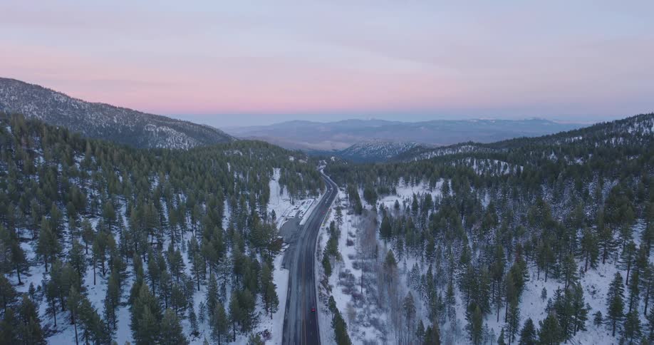 Light traffic moves on a snow covered forested mountain road with mountains in the distance