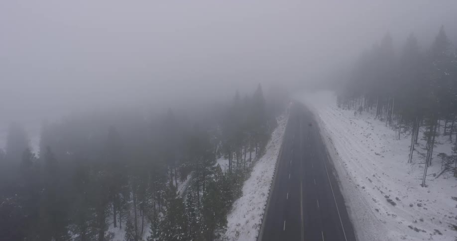 A couple of cars pass by on a mountain pass highway surrounded by snow and pine trees and fog becoming thicker just as it begins to get dark