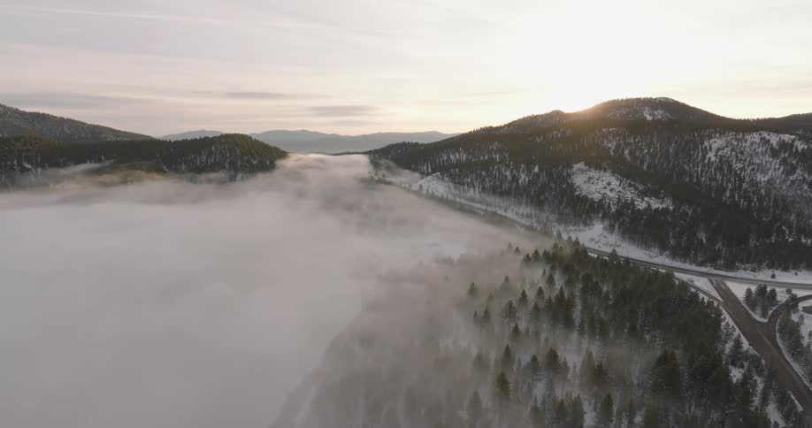 Aerial of a fogged covered pine tree forest next to a mountain pass highway lined with snow and the sun glaring from behind hill peak in the background