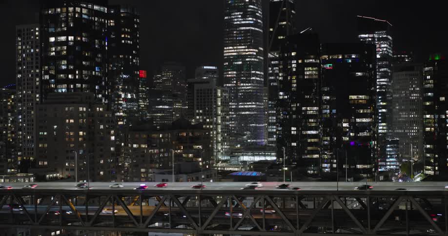 Cars drive across a bridge in two directions in front of a city full of brightly lit windows at night