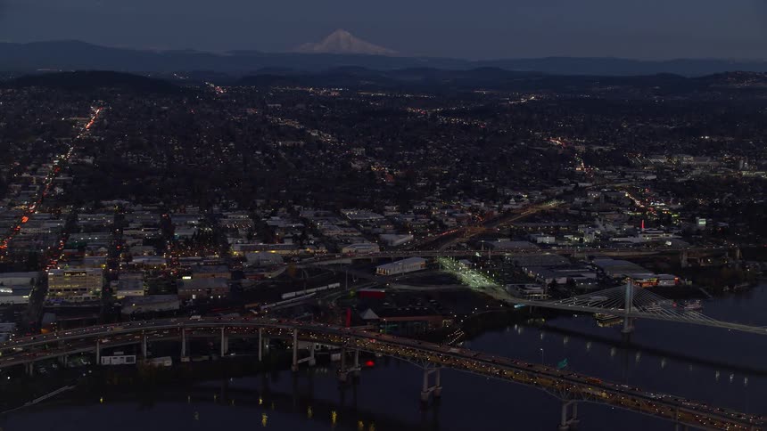 Traffic crosses dual level causeway near waterfront of city bordering white topped mountain after dusk