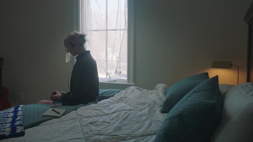 Blonde woman with headphones and a black shirt sits on bed