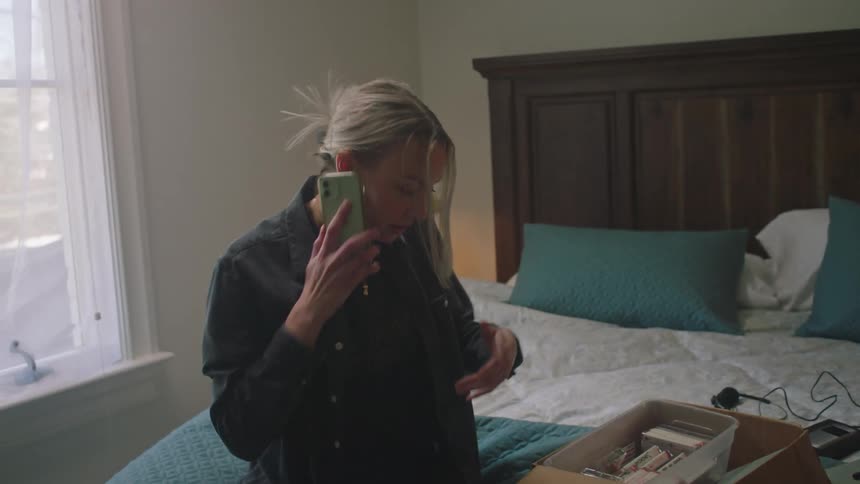 Lady sits on the bed to check through a carton of cassette tapes while making call