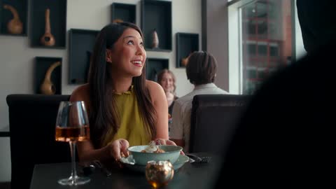 A woman in a restaurant holding a glass of wine is served food and is happily surprised by the dish