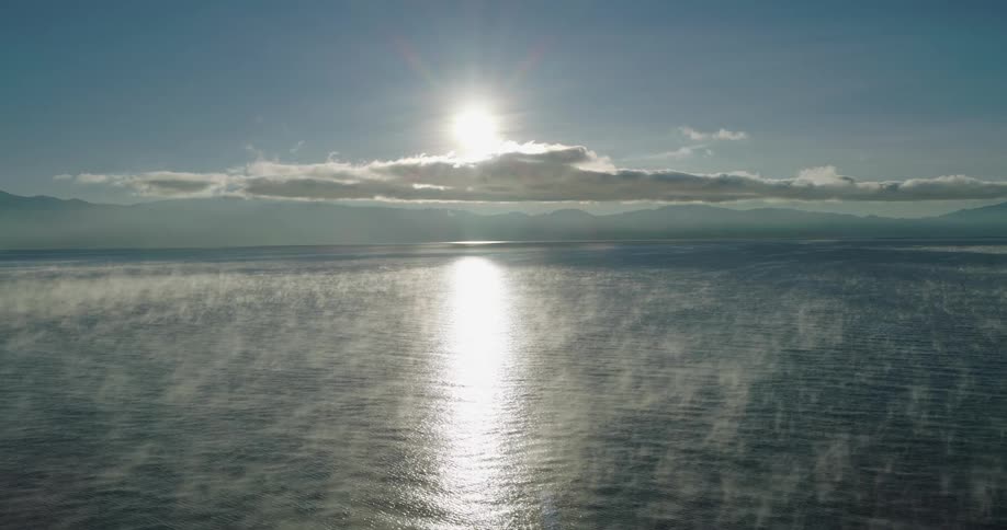 The sun shines above clouds over mountains reflecting on the water along the coast