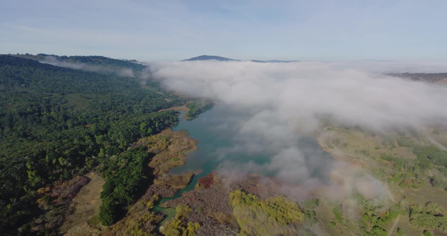 Aerial of a lake covered in clouds of fog and a lush green hillside on a bright day