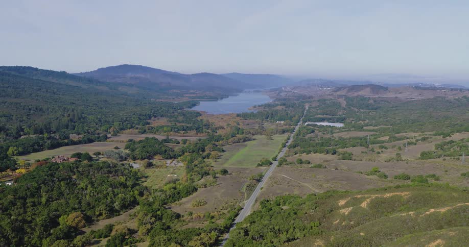 Aerial panoramic of a road through a rural valley landscape with a lake and mountains in the background on a sunny day