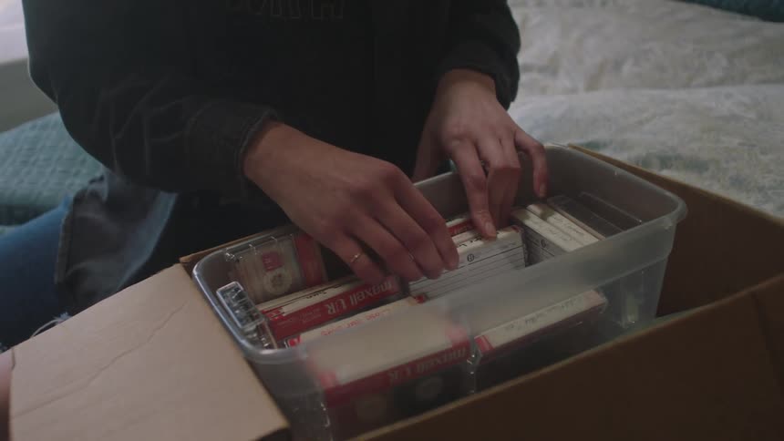 Woman sitting on the bed talking on the phone and looking at tapes in a box