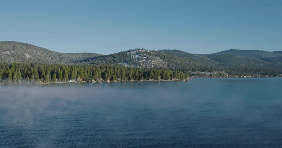 Thin fog on top of a lake surrounded by a forest of pine trees with hills and peaks in the background and a blue sky on a sunny day