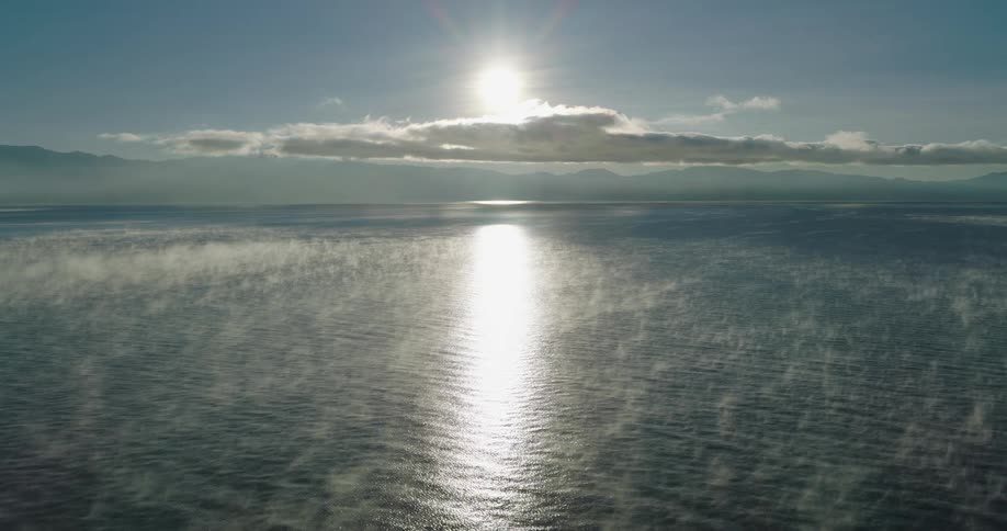 The sun shines above clouds over mountains reflecting on the water along the coast