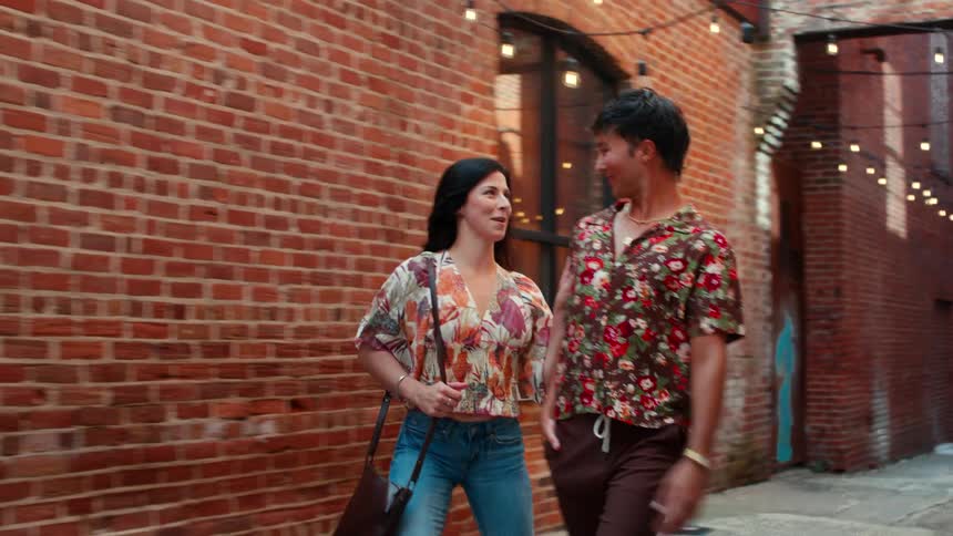 Man and woman walk down an alley with overhead string lights while talking and laughing together