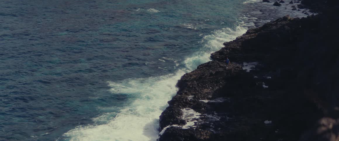 Waves roll into shore and splash against rocks along the coast as a man walks on a rock overlooking the water