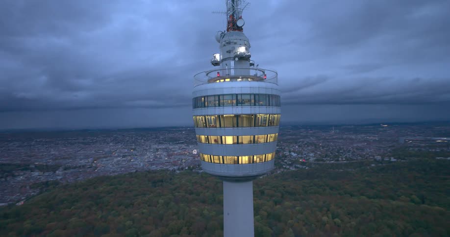 A large white tower with lights inside and behind it is a large city and forests