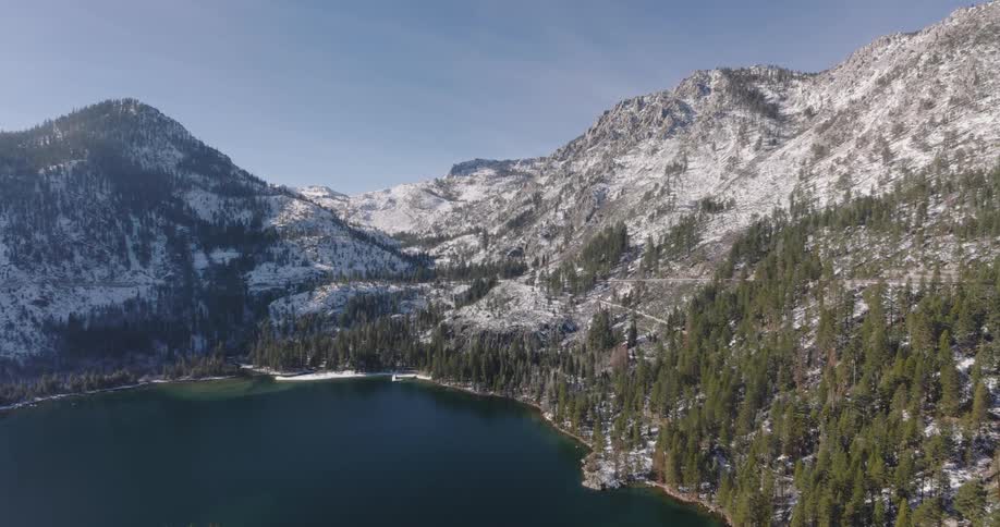 Low aerial view of a lake beside snow dusted pine tree covered slopes with rocky mountain peaks during the day
