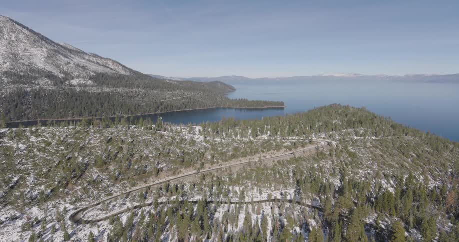 Aerial view of a winding road on a coastal hillside dusted with snow a pine trees beside a lake with a mountain peak nearby during the day