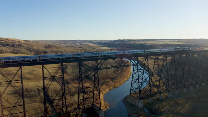 A passenger train travels on a trestle bridge over a stream as the sun shines brightly in the distance