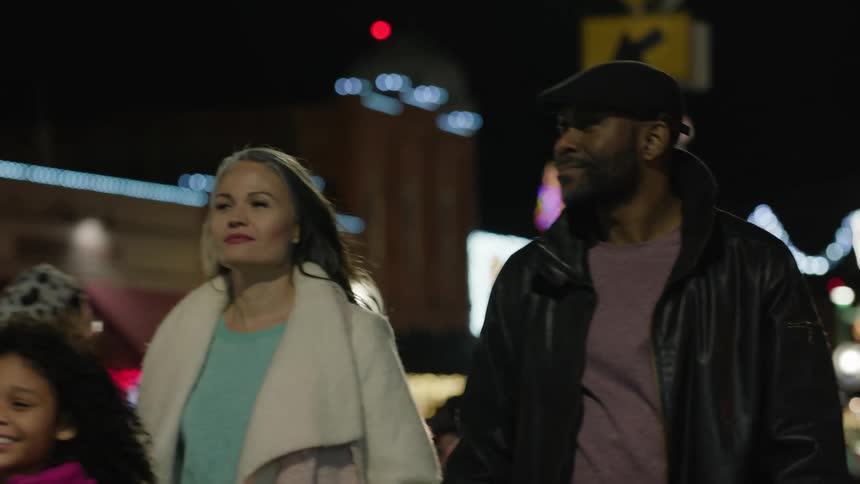 Parents walking hand in hand with their young daughter leading the way on a sidewalk at night in the city