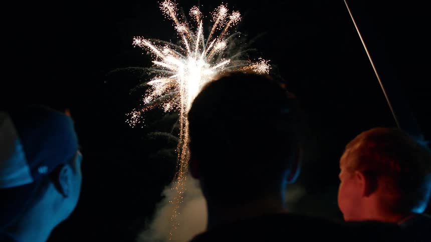 Three people including a father holding a son look up at fireworks exploding at night in the sky