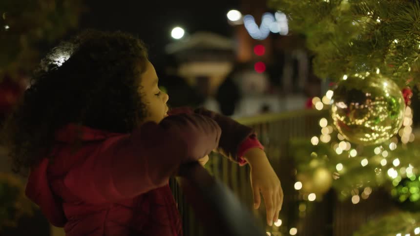 Girl reaches over fence to touch large golden ornament on outdoor christmas tree at night