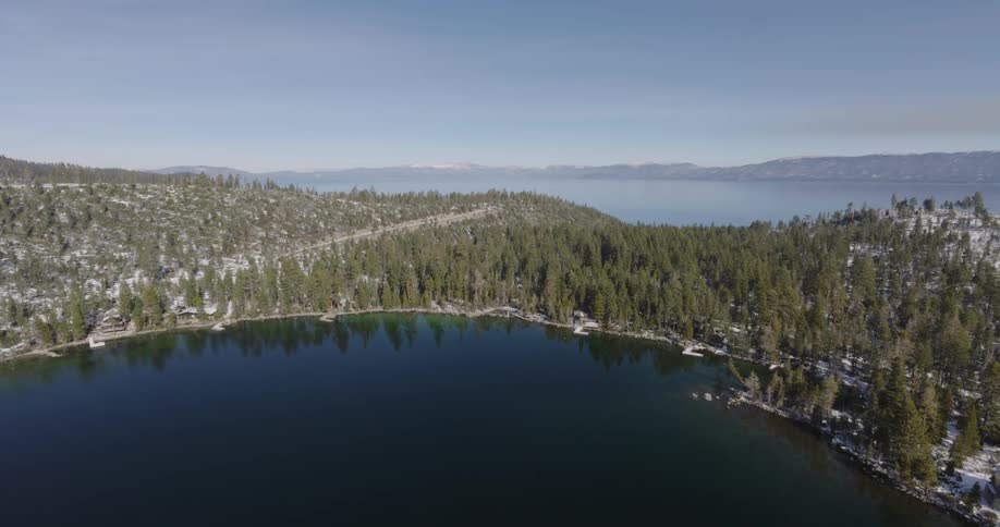 Aerial over a dark lake a thin strip of land covered in snow and pine trees in the middle and another body of water under slight fog on a bright day