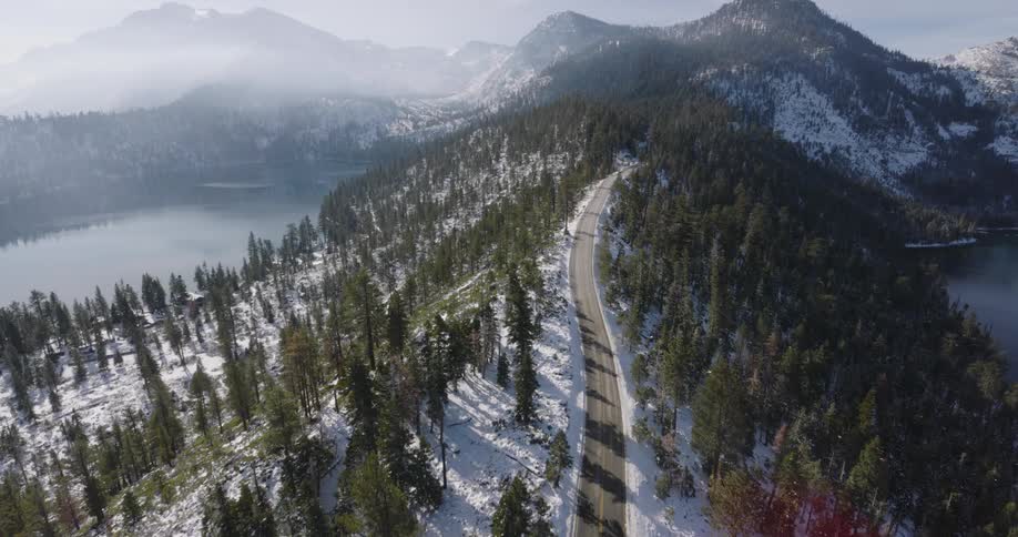 An automobile travels on a road across the top of a snow covered forested mountain with lakes on each side