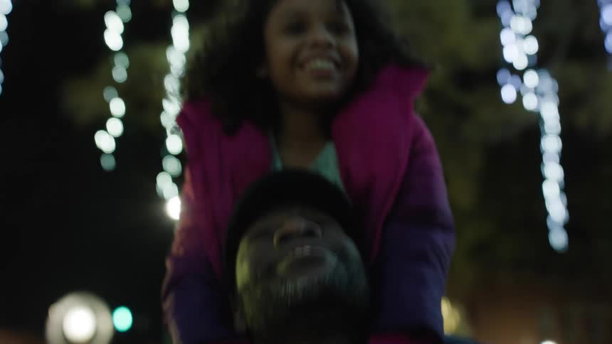 Daughter sits on her fathers shoulder while admiring christmas decorations outdoor at night