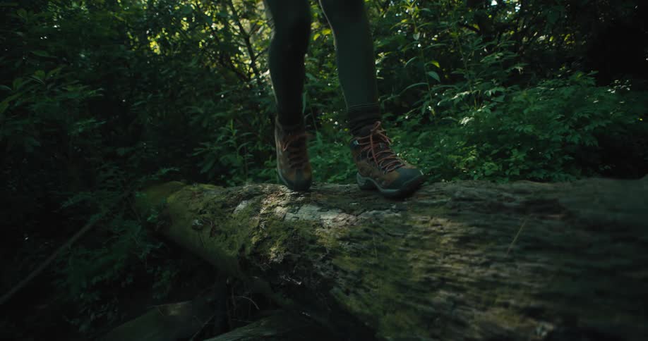 Woman in orange crop top and green pants walks across a mossy log in the forest