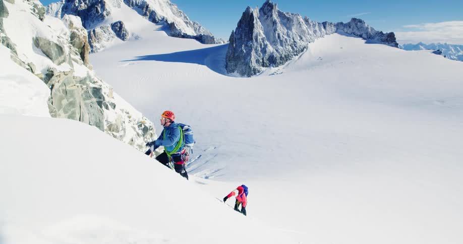 Woman and man climb a steep mountain covered in powdery snow on a bright blue day