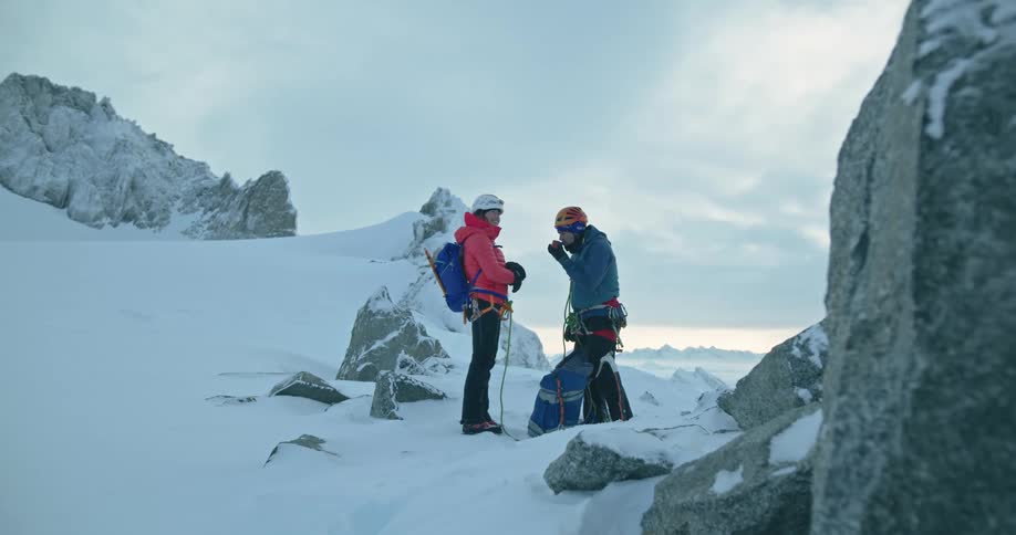 Two climbers tied together with a rope stand on snowy mountain and drink from a shared cup