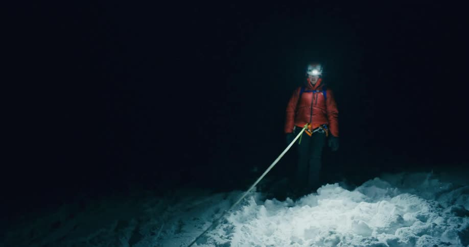 Male and female mountain climbers wearing winter weather gear and headlamps and tied together with rope walk through snow at night